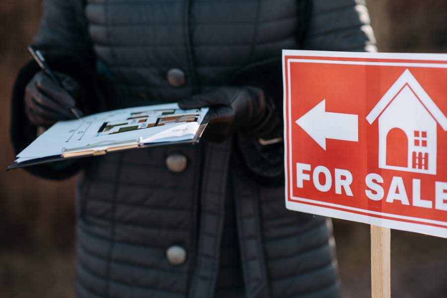 Person in black jacket beside a house for sale sign