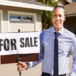 a real estate agent in blue shirt holding a 'FOR SALE' sign