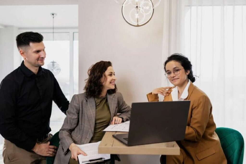 Realtor showing something on her gray laptop to a woman who is sitting down next from her holding white legal papers while her husband is standing close