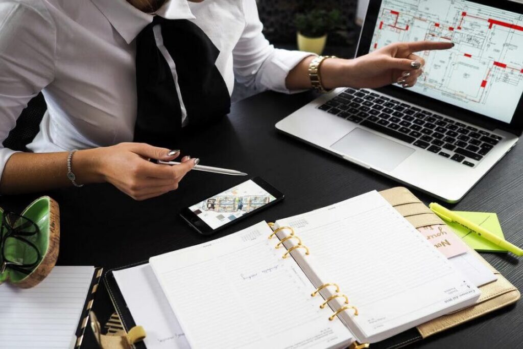 Realtor wearing a white dress shirt pointing to a house blueprint on her silver laptop while her notebook and phone are laid open on her table