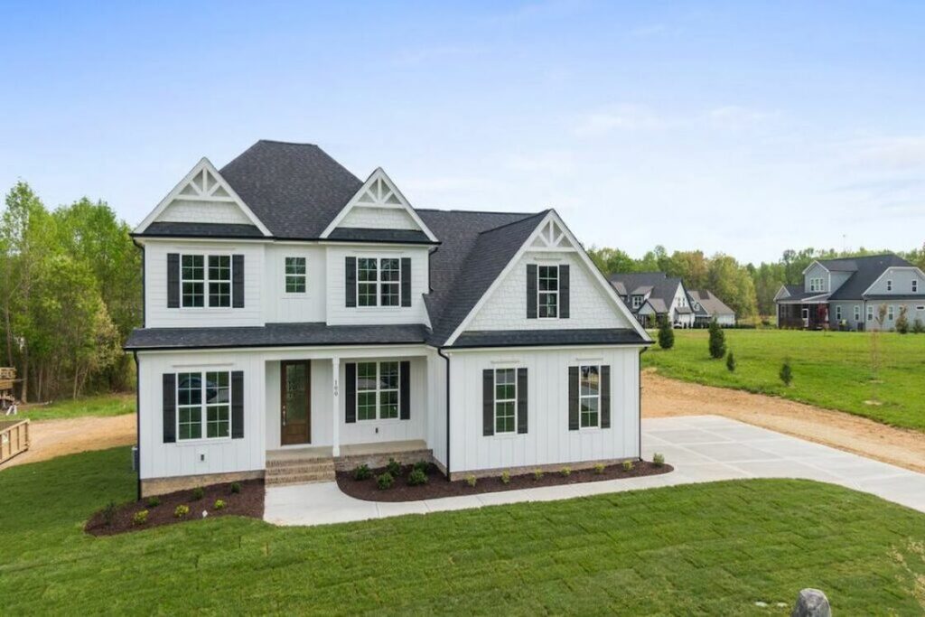 Drone shot of a house with gray roofing and white painted walls and green lawn, with the nearby houses also visible in the photo