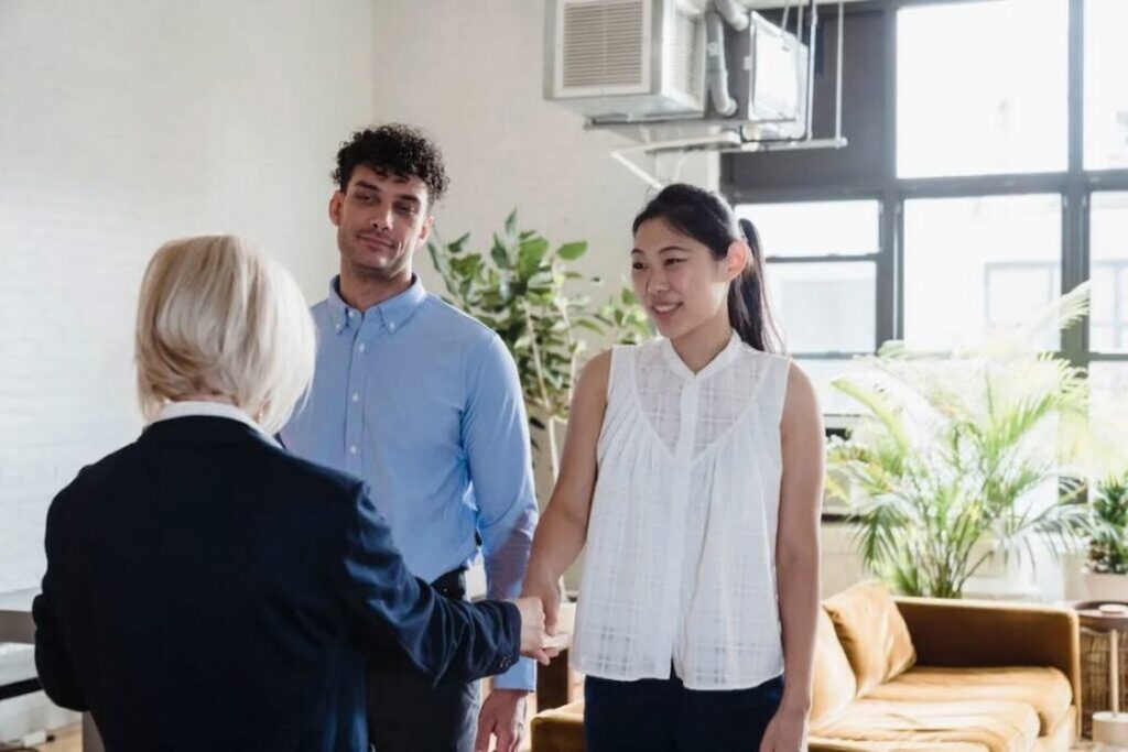 Woman wearing a white sleeveless top shaking hands with a real estate agent wearing a dark blue suit while her husband looks on