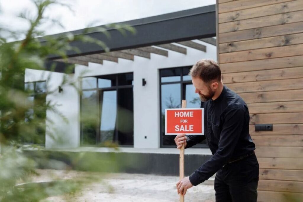 Man wearing a dark blue longsleeved dress shirt and black jeans installing a red and white "home for sale" sign