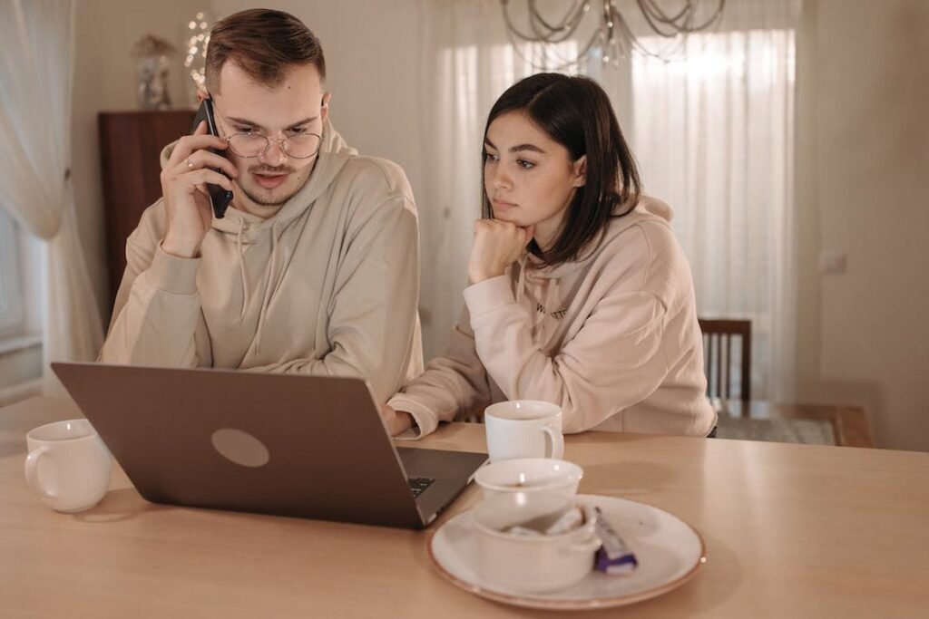 Man talking on his black smartphone while he and his wife are looking at their gray laptop