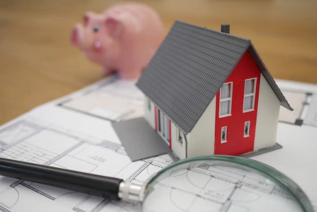 Miniature house on top of a floor plan and beside a magnifying glass and piggy bank