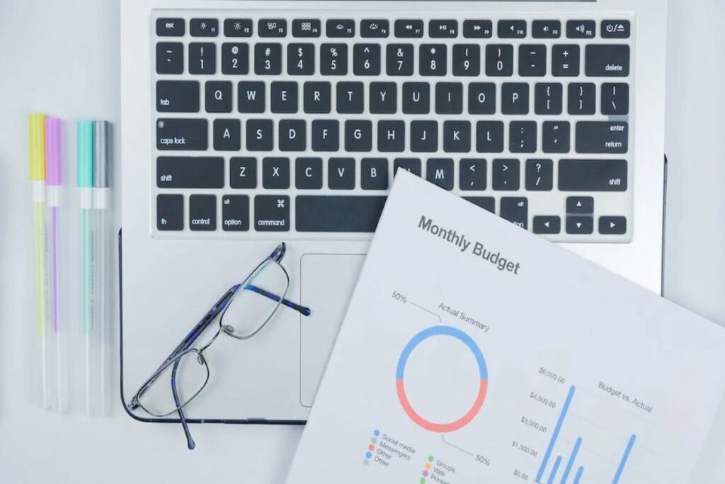 Printed copy of a monthly budget report placed on top of a silver laptop and beside a reading glass and pens